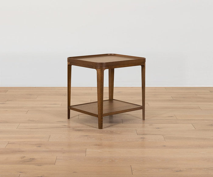The Aden End Table, a compact square piece crafted from solid oak with slender legs and a lower shelf, is set on a light hardwood floor against a plain white wall.