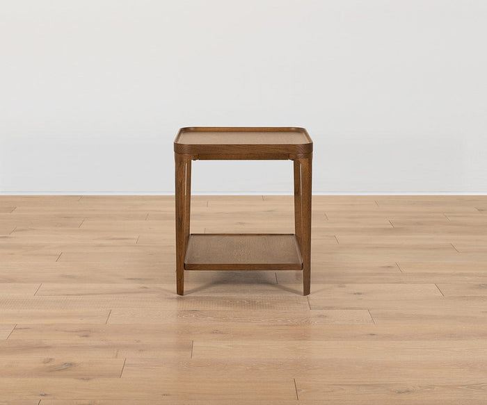 The Aden End Table, part of the Aden Collection, is a compact wooden side table with a square top and lower shelf. Supported by four legs, it stands on a light wood floor against a simple white wall, offering a blend of simplicity and elegance in true mid-century style.
