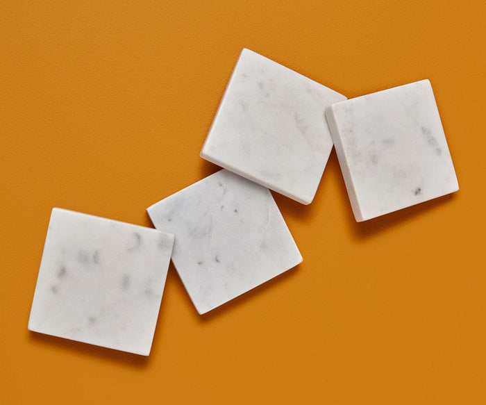 Four Adia Square Marble Coasters – 4 Piece, crafted from white marble and featuring subtle gray veining, are handcrafted in India and arranged in a loose diagonal line on an orange surface.