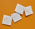 Four Adia Square Marble Coasters – 4 Piece, crafted from white marble and featuring subtle gray veining, are handcrafted in India and arranged in a loose diagonal line on an orange surface.