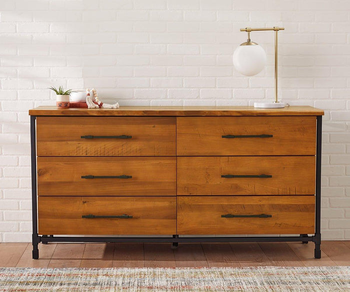 A wooden dresser with six drawers features black metal handles and black legs. On top sits a potted plant, a seashell, a small decoration, and the Aerlig Table Lamp, which brings modern elegance to the setup. The scene is set against a white brick wall with a colorful rug in the background.