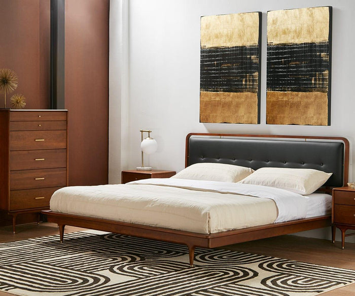 A modern bedroom features a wooden bed dressed in white bedding, accented with a dark headboard and complemented by matching nightstands and a dresser. A geometric rug grounds the space, while the Aerlig Table Lamp sits beside the bed, casting a warm, inviting glow. Two abstract gold and black paintings on the wall enhance the contemporary aesthetic.