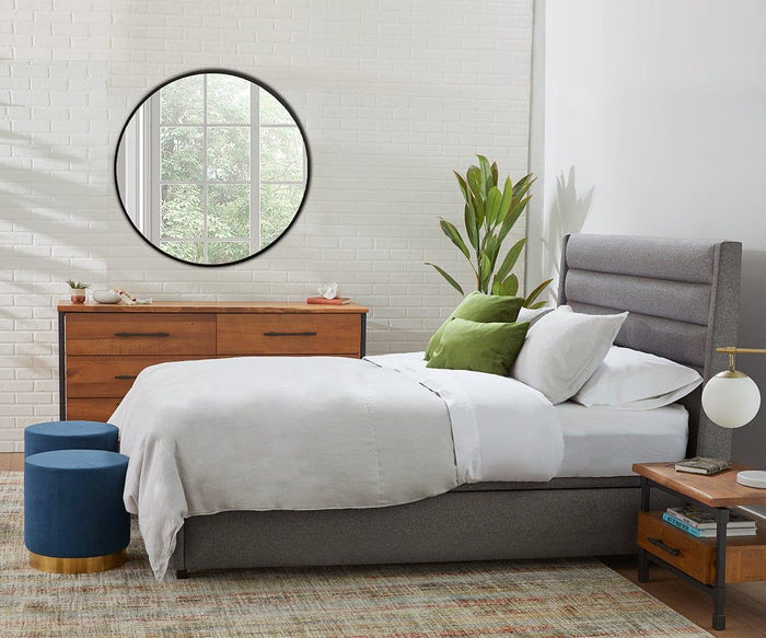 A modern bedroom features a gray upholstered bed with white bedding and green pillows, complemented by a wooden dresser topped with a round mirror. Two blue ottomans sit alongside a plant, while the bedside table is adorned with the Aerlig Table Lamp. A large window reveals lush greenery outside.