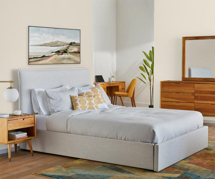A modern bedroom features a gray upholstered bed, wooden nightstand, dresser, desk, chair, a potted plant, and a landscape painting above the bed. Light-toned bedding and walls create an airy feel, complemented by a colorful rug on the floor. The Aerlig Table Lamp sits on the nightstand, adding both style and warm illumination to the space. A mirror completes the room’s contemporary look.