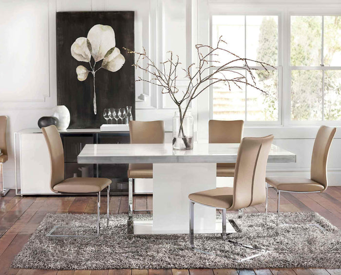 Modern dining area with a white table, Alma Dining Chairs in beige, a gray rug, a vase with branches, and a large floral artwork on the wall. Sunlight streams in through expansive windows, creating a bright, elegant atmosphere that epitomizes contemporary style.