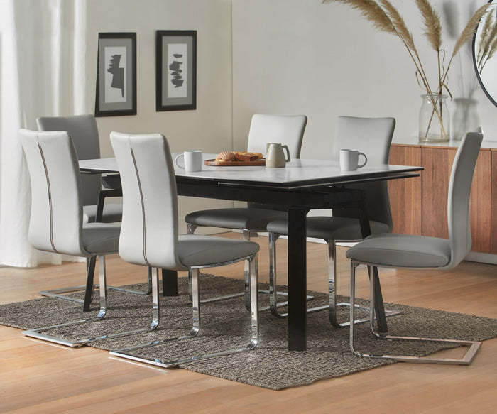 A modern dining area features a rectangular table surrounded by six Alma Dining Chairs in gray, accompanied by mugs, pastries, and a teapot. The space is decorated with framed artwork, vases filled with pampas grass, and a cabinet placed on a woven rug. Sunlight streams through white curtains, enhancing the contemporary ambiance of the room.