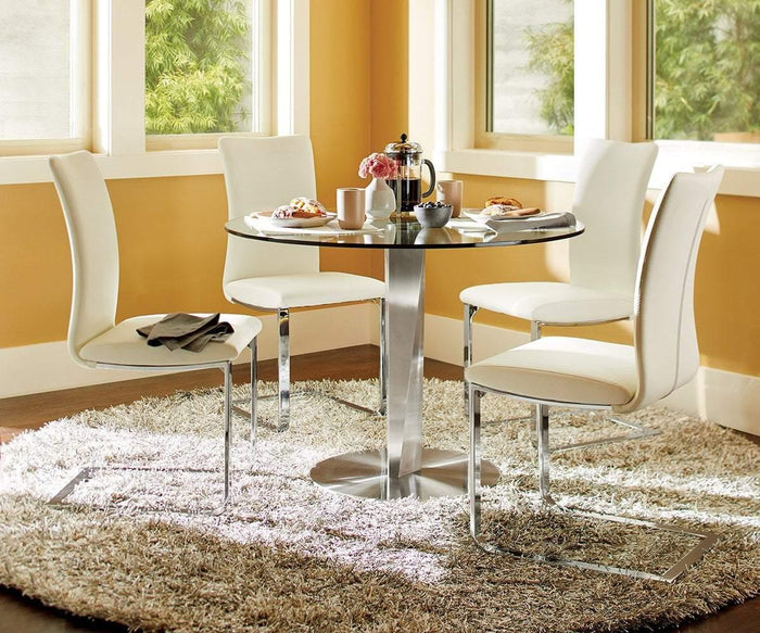 A round glass table with four Alma Dining Chairs sits on a circular rug in a bright dining area. The table is set with a French press, mugs, plates of pastries, and neatly folded napkins. Large windows let in abundant natural light, highlighting the contemporary style of the space.
