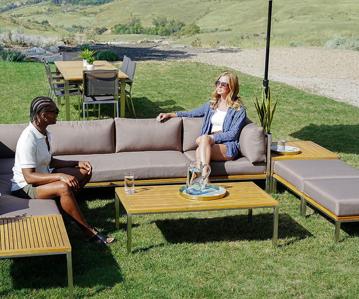 Two people sit on a modern outdoor patio sofa, talking and relaxing. An Ambra Tray with a gold rim holds a water pitcher and glasses on the table between them, adding an elegant accent to the scene. There’s a grassy lawn and dining table set in the background.