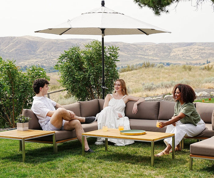 Three people relax on an outdoor patio set under a large umbrella, with hills in the background. Two sit on a couch, and one sits on the edge, all smiling with drinks resting on the Ambra Tray—an elegant enamel piece with a gold rim—accenting the wooden table.