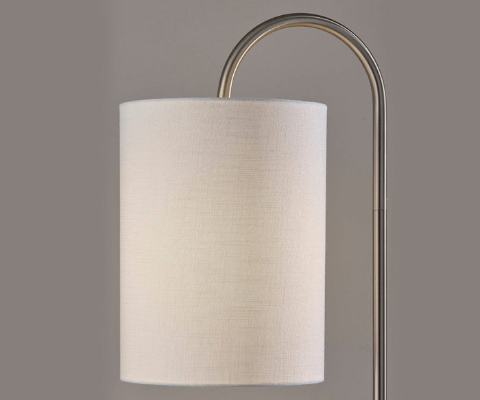 A close-up of the Amira Shelf Floor Lamp highlights its sleek modern silhouette, featuring a curved metal arm, a cylindrical white fabric lampshade, and an elegant brushed steel finish against a plain gray background.