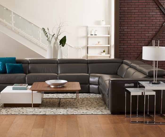 Modern living room featuring a gray leather sectional sofa, wooden coffee table, white side tables, and blue cushions; accentuated by decorative vases and the soft illumination of the Anderson Table Lamp with its elegant linen drum shade. A brick wall and staircase serve as a stylish backdrop.