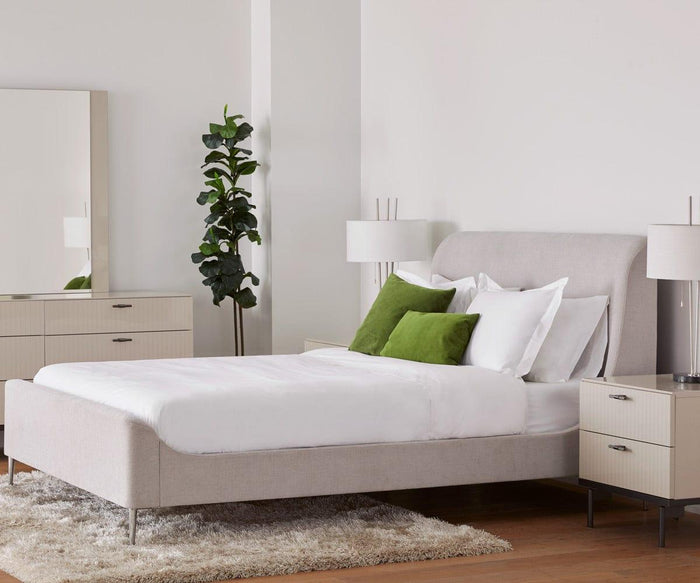 A modern bedroom featuring a beige upholstered bed with white bedding and green accent pillows, a dresser with mirror, nightstands, two Anderson Table Lamps with elegant linen drum shades, a potted plant, and a light rug on wooden flooring.