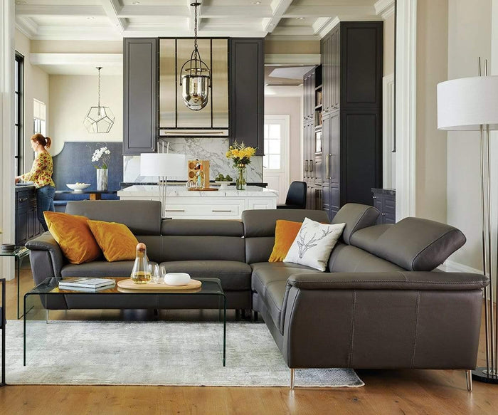 A modern living room features a gray sectional sofa accented with orange and yellow pillows, a glass coffee table, and an open kitchen in the background where a woman stands at the counter. The space is softly illuminated by the Anderson Table Lamp with its linen drum shade, enhancing the warm and inviting atmosphere.