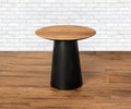 The Andor End Table, featuring a natural teak round wooden top and a black cone-shaped base, stands on a wooden floor before a white brick wall, evoking the classic elegance of mid-century design.