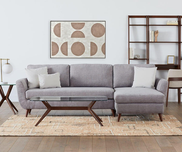 A modern living room with a gray sectional sofa, two white pillows, a glass coffee table, geometric wall art, a bookshelf, and an Ardal End Table with a lamp. The space features a beige patterned rug on a wooden floor and is accented by European beech wood touches for added warmth.