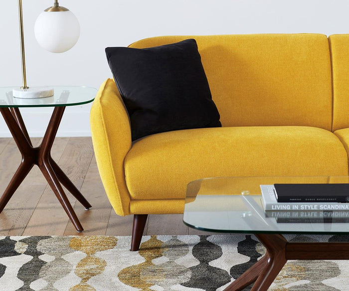 A modern living room with a yellow sofa, black pillow, wooden side and coffee tables with glass tops, a lamp, and a patterned rug in neutral tones. The Ardal End Table in European beech wood adds elegance next to the sofa, while a book sits neatly on the coffee table.
