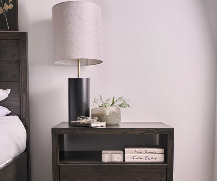 A dark wood nightstand with the Ash Table Lamp—a modern rustic lamp featuring a dark wood base and a black and white shade—alongside a small plant in a white textured vase, a pair of glasses, two books, and stacked decorative books labeled Paris, France and London, England.