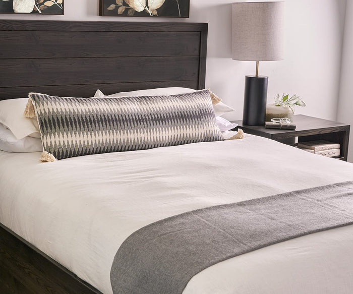 A neatly made bed with white bedding, a gray throw, a long patterned pillow, and a dark wood headboard is complemented by a bedside table featuring the Ash Table Lamp, books, and a small plant.