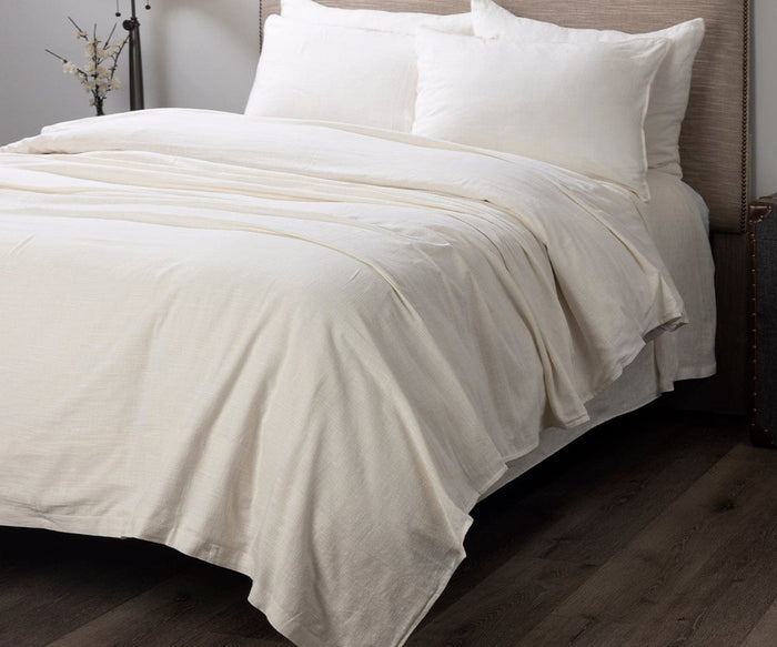 A neatly made bed features the Baker Duvet Cover in Ivory A04 and matching pillows, set against a beige headboard. The room has dark wood flooring and a small decorative vase in the corner.