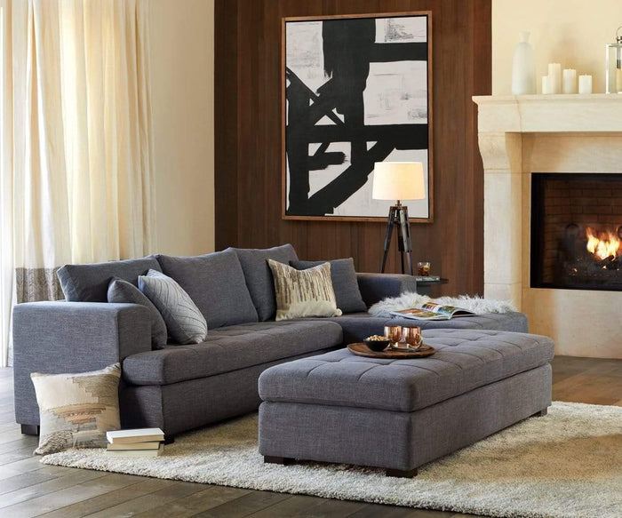 A modern living room featuring a gray sectional sofa and ottoman, neutral decor, and the captivating Balkon Abstract Graphic Wall Art as the centerpiece—a striking black-and-white painting displayed above a stone fireplace with a cozy fire burning. Light filters gently through sheer curtains, softly illuminating the space accented by a floor lamp and understated architectural details.