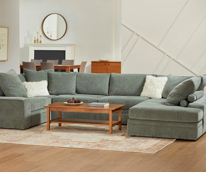 A spacious living room with a large gray sectional sofa, two white pillows, a wooden coffee table, and a light rug. In the background are a dining table, chairs, a fireplace, and the Baptisia 35″ Round Mirror with its elegant wood frame displayed on the wall—an inviting showcase of modern artistry.