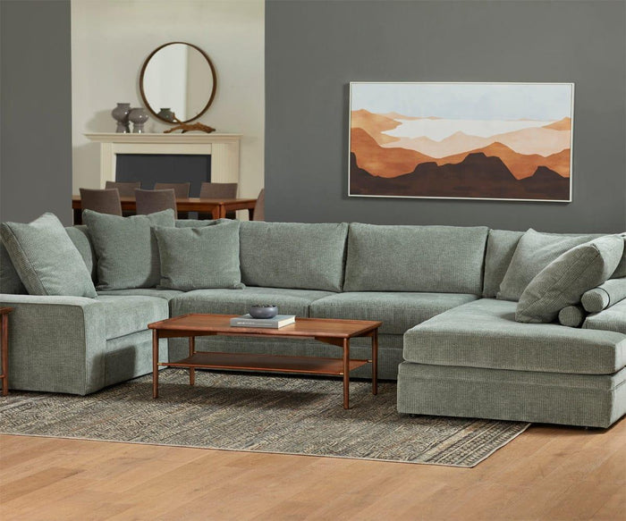 A gray sectional sofa with a wood frame and a wooden coffee table sits on a patterned rug. A landscape painting and the Baptisia 35″ Round Mirror decorate the gray wall, highlighting the room’s modern artistry. The background includes a dining table and fireplace.