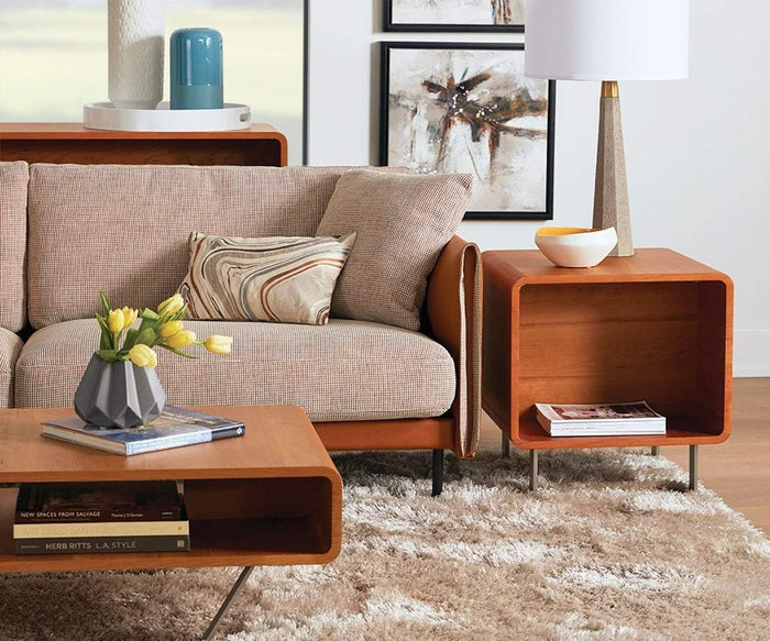 A modern living room features a beige sofa and a wooden coffee table, with the elegant Baptisia End Table in cherry finish serving as an eye-catching accent. The end table is topped with a stylish lamp and decorative bowl, while abstract wall art and a striking cubist sculpture add artistic flair to the space. A vase of flowers and a plush beige shag rug on the wooden floor complete the inviting look.