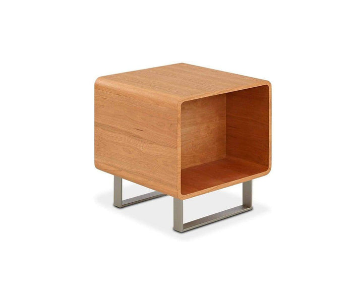 The Baptisia End Table is a modern wooden piece with rounded edges and an open cubby, supported by two metal legs. Its minimalist design, reminiscent of a cubist art sculpture, highlights the wood’s warm cherry finish.