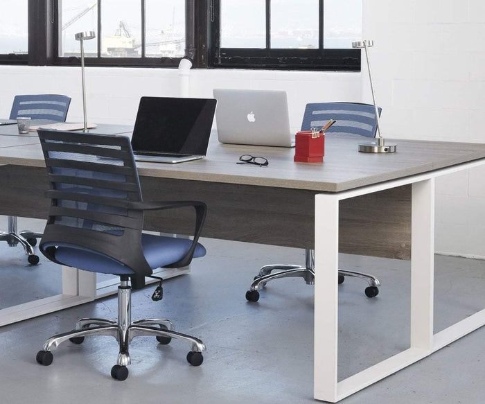 Modern office workspace with two laptops, a notebook, glasses, pens, and desk lamps on a shared wooden table. Two Barrier Desk Chairs with mesh backrests are positioned at the desk near large windows.