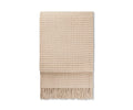 The Basket Throw, shown folded on a white background, features a beige woven design crafted from recycled alpaca wool with a subtle grid-like texture and long fringe along the bottom edge. Cozy and stylish, this throw is ideal for bringing warmth and elegance to any room.