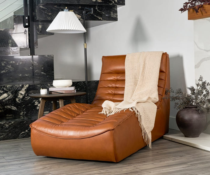 A modern brown leather chaise lounge is draped with the Basket Throw, a cozy blanket made from recycled alpaca wool, beside a small round table and floor lamp in a room featuring marble stairs and wood flooring. Decorative vases with branches complete the inviting, fair trade-inspired setting.
