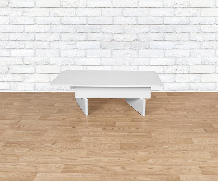 The Batia Coffee Table, featuring geometric legs, sits low on a light wood floor in front of a white brick wall, infusing the space with contemporary style.