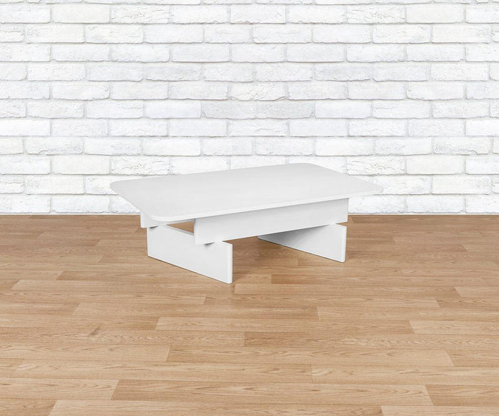 The Batia Coffee Table, featuring a small modern design with geometric legs, sits on a light wood floor in front of a white brick wall.