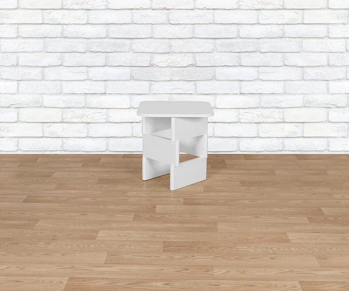 The Batia Side Table, with its small white modern design and open shelves, sits on a light wood floor in front of a white brick wall, adding a sleek and functional touch to the contemporary space.