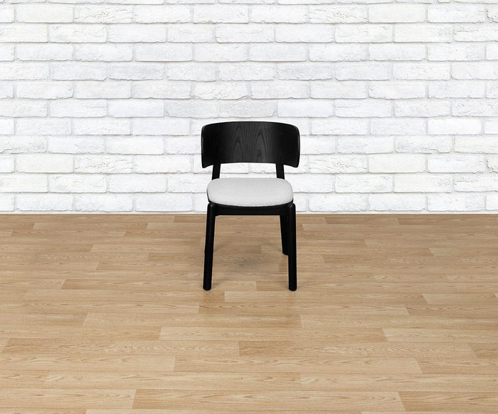 A contemporary Beau Chair in black wood with an upholstered seat rests on a light wood floor in front of a white brick wall.