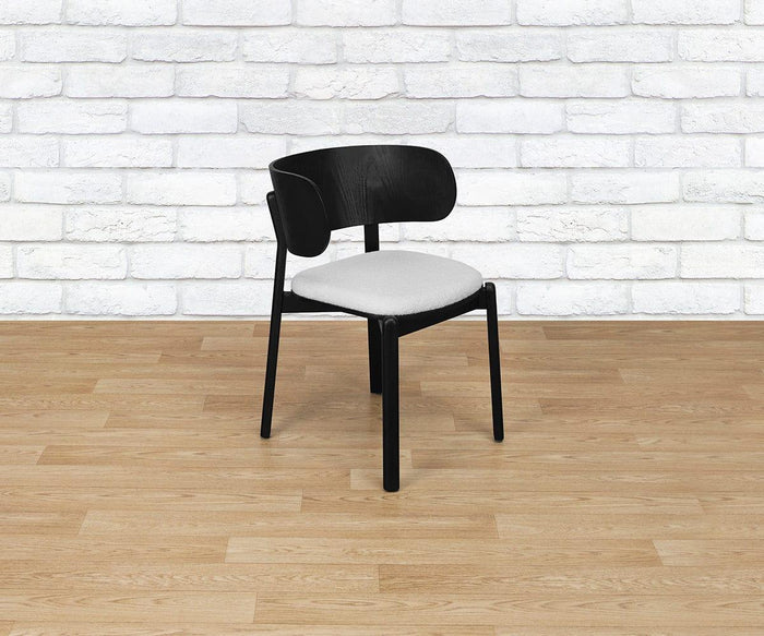 A Beau Chair featuring a modern black wooden frame, curved backrest, and upholstered light gray seat is placed on a light wood floor in front of a white brick wall.