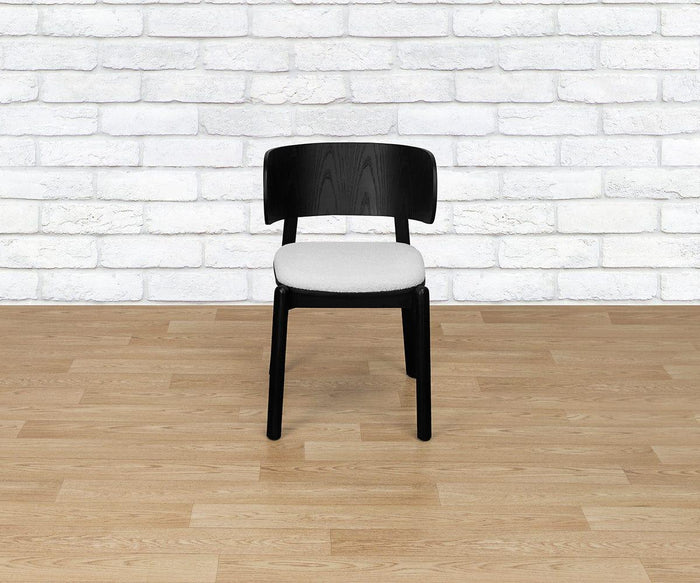 The Beau Chair, featuring a black wooden frame, a curved backrest, and a light gray upholstered seat, is placed on a light wood floor in front of a white brick wall.