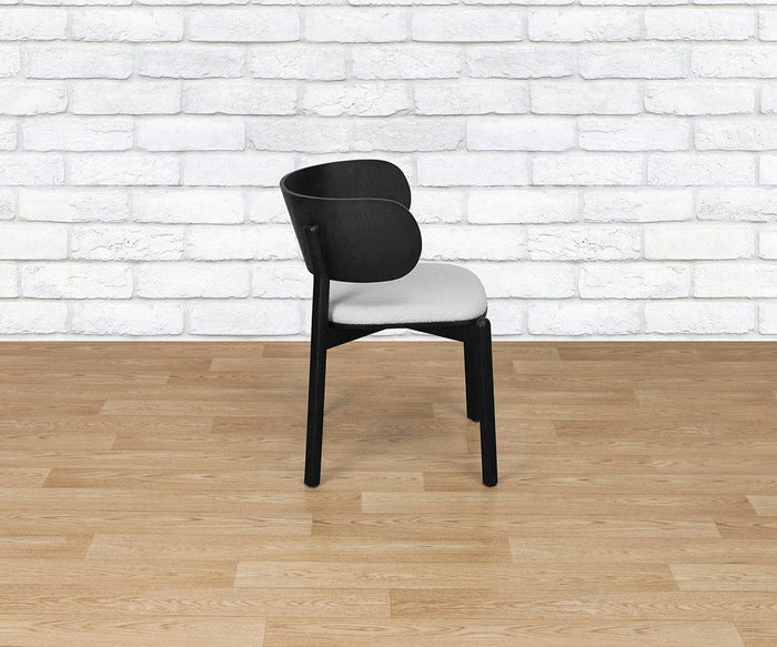 The Beau Chair, a modern black wooden dining chair with an upholstered seat, is placed on a wooden floor in front of a white brick wall.