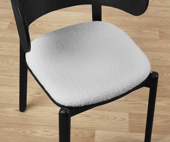 A black wooden Beau Chair with an upholstered seat featuring a soft, white textured cushion rests on a light wood floor. The photograph is taken from an overhead angle, highlighting the seat.