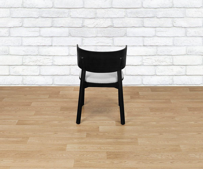 A Beau Chair in black wood with an upholstered seat is positioned facing away from the camera, set atop a light wood floor with a white brick wall as the backdrop.