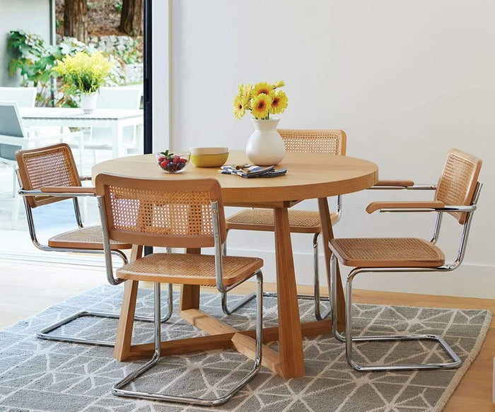 A round wooden dining table with four Bendt Dining Chairs featuring chrome frames and cane seats sits on a patterned rug. A vase with yellow flowers, a bowl, and a plate of fruit are arranged on the table. An outdoor patio is visible in the background.