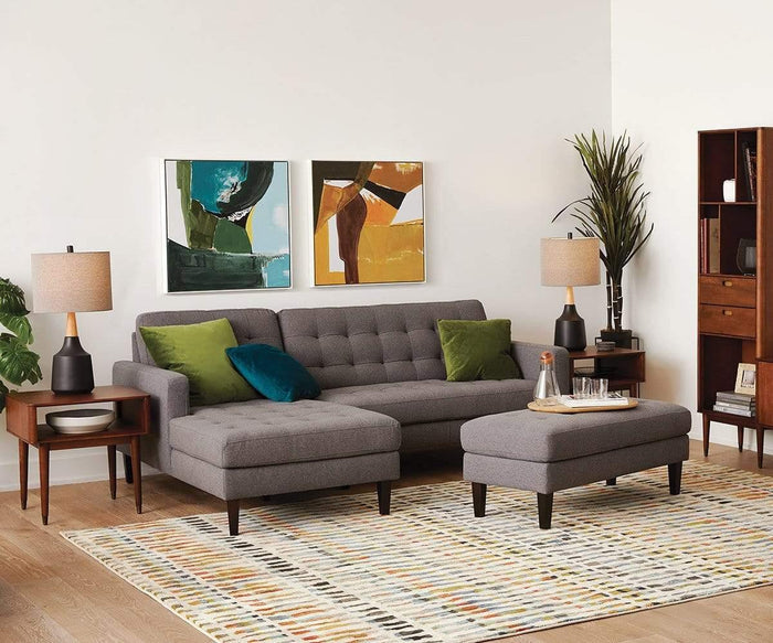 Modern living room with a gray sectional sofa, green pillows, two abstract paintings, side tables topped with Bergset Table Lamps—a stylish lighting choice featuring a black ceramic base and grey fabric drum shade—an ottoman, patterned rug, potted plant, and wooden bookshelf set against white walls.