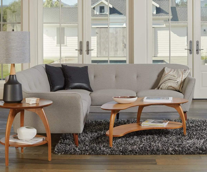 Modern living room with a gray sectional sofa, three cushions, two wooden tables, a Bergset Table Lamp featuring a grey fabric drum shade on a black ceramic base, a bowl, and magazines atop a textured gray rug. Large windows and glass doors reveal a house and trees outside.