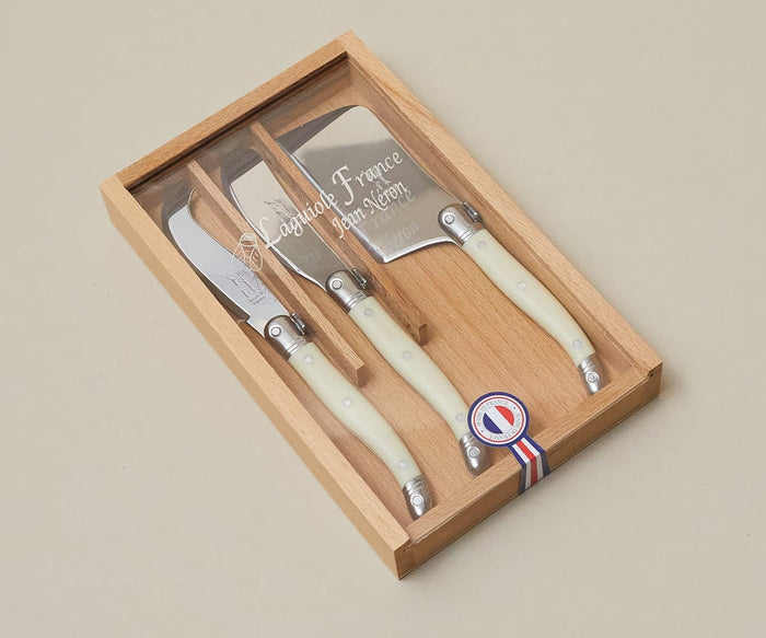 A wooden box holds the Brava Ivory Cheese Set, featuring white-handled cheese knives with stainless steel blades, labeled Laguiole Jean Néron—renowned for its fine French cutlery—displayed on a beige surface.