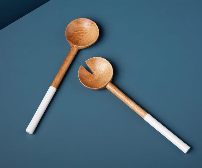 The Bright Serving Set, crafted from mango wood, features two salad servers—one solid and one with a forked notch—each accented with food-safe enamel and white handles. Placed on a blue surface, this handcrafted set adds an elegant, functional touch to your table.