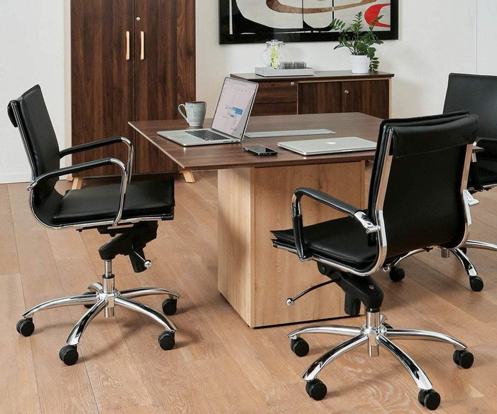 Modern office featuring the Brock Low Back Office Chair and black swivel chairs arranged around a wooden table, accompanied by an open laptop, a closed laptop, a notebook, and a coffee mug. A plant and artwork add accents in the background atop a wood floor.