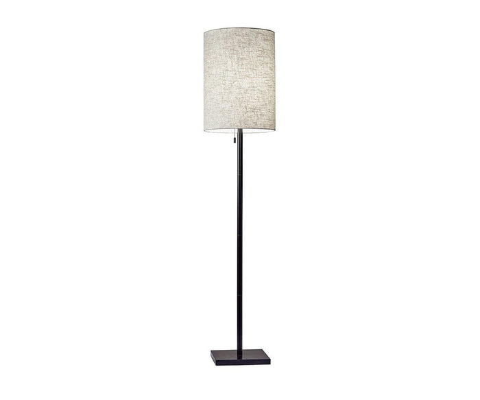 The Cara Floor Lamp features a tall, slim dark bronze metal base and a cylindrical beige fabric shade, standing on a flat square base against a white background.