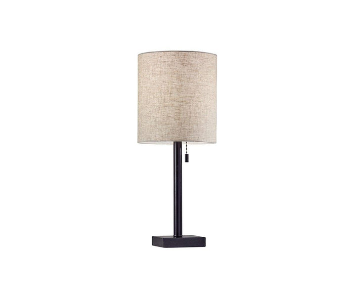 The Cara Table Lamp showcases a modern design with a cylindrical beige fabric shade, a slim black metal stand, and a sleek rectangular metal base. A small pull chain hangs just beneath the shade. The background is plain white.