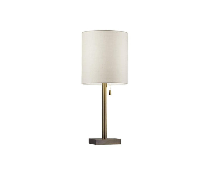 The Cara Table Lamp features a modern design with a cylindrical white fabric shade, a slender brass-finished metal stem, and a rectangular dark metal base. A pull chain switch hangs just below the shade.