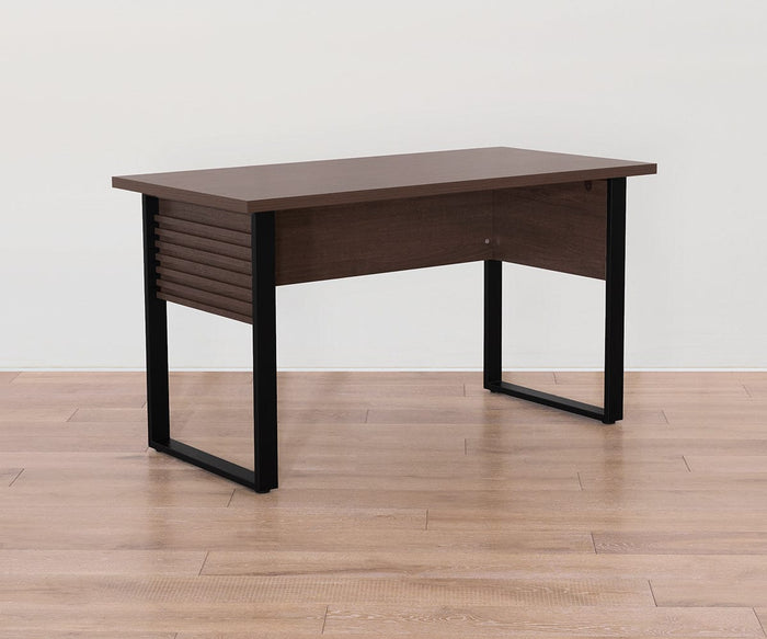 A modern Cary Desk featuring a 55″ rectangular dark wood melamine top, panel sides, and black metal legs, positioned on a light wood floor against a plain white wall.